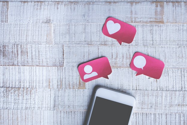 Mobile phone on tables surrounded by post it notes cut into the shape of word bubbles containing heart, person, and comment emojis.